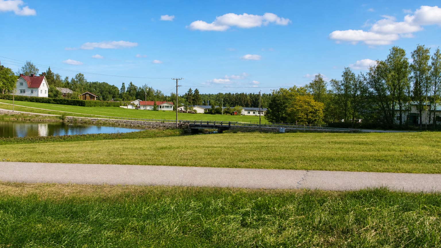 Grassy meadow and footpath by a small pond with a footbridge and red-roofed wooden houses in rural Nurmijärvi, Finland