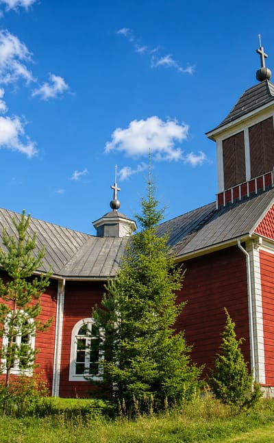 Red wooden church with a white-trimmed bell tower and crosses on a hill in Pukkila, Finland.