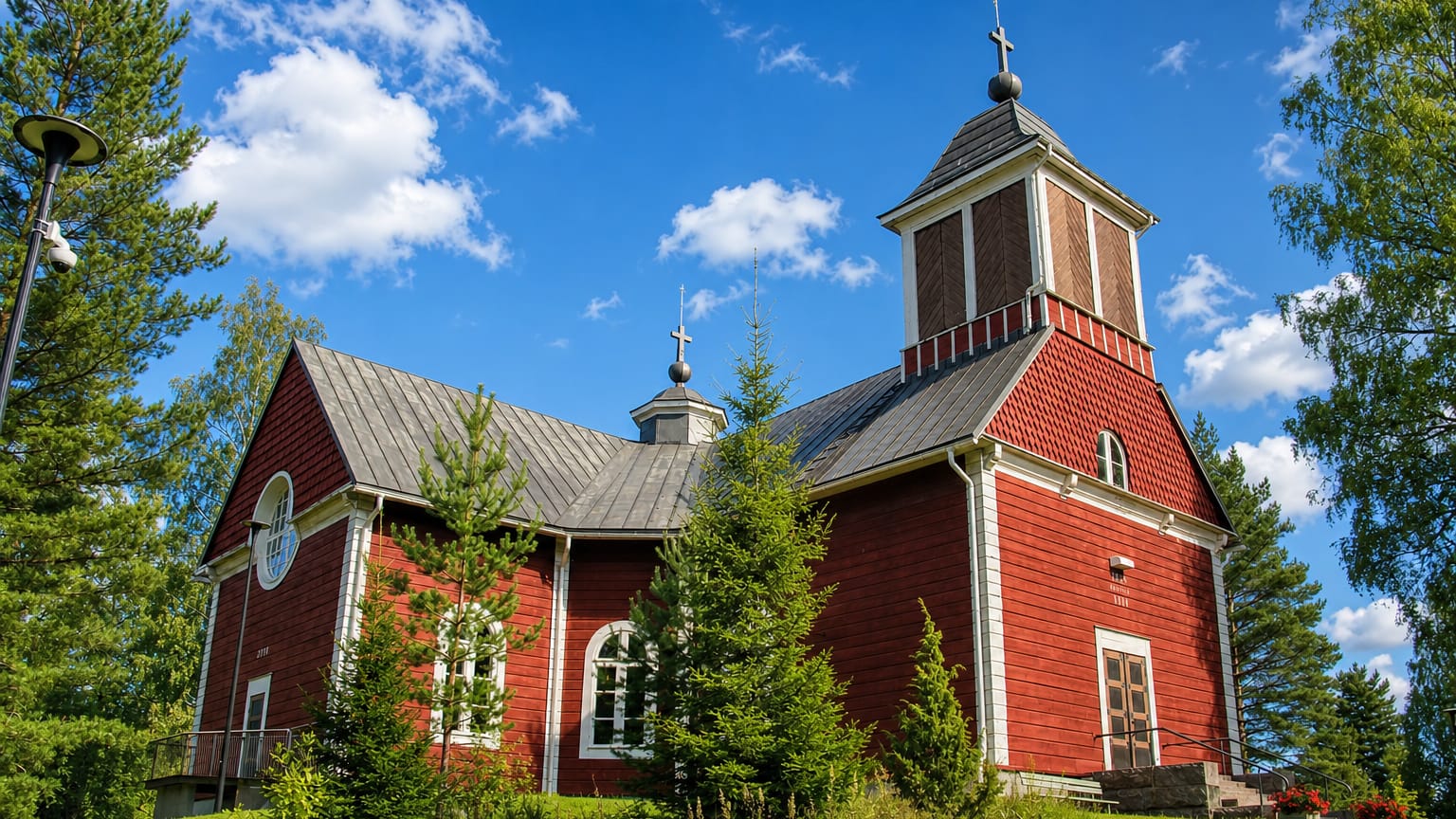 Red wooden church with a white-trimmed bell tower and crosses on a hill in Pukkila, Finland.