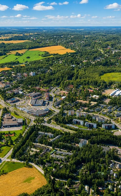 Aerial view of Tuusula, Finland, with a low-rise town centre among green fields, forest and a lake under blue sky.