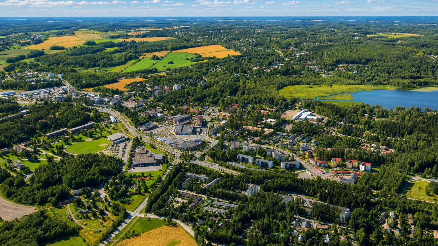 Aerial view of Tuusula, Finland, with a low-rise town centre among green fields, forest and a lake under blue sky.