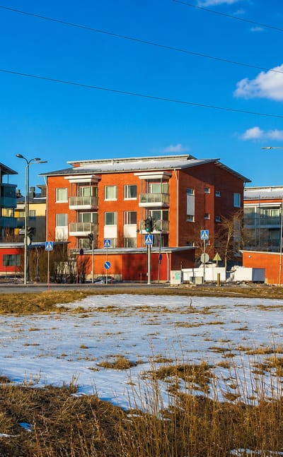 Modern red and yellow apartment blocks in Pakkala, Vantaa, beside a road with patchy snow under a blue winter sky.