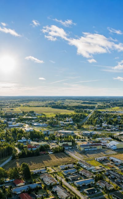 Aerial daytime view of Aura, Finland, with low buildings spread among green fields and forests under a bright sunlit sky.