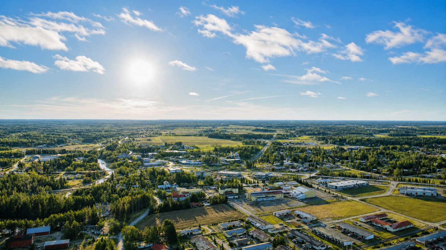 Aerial daytime view of Aura, Finland, with low buildings spread among green fields and forests under a bright sunlit sky.