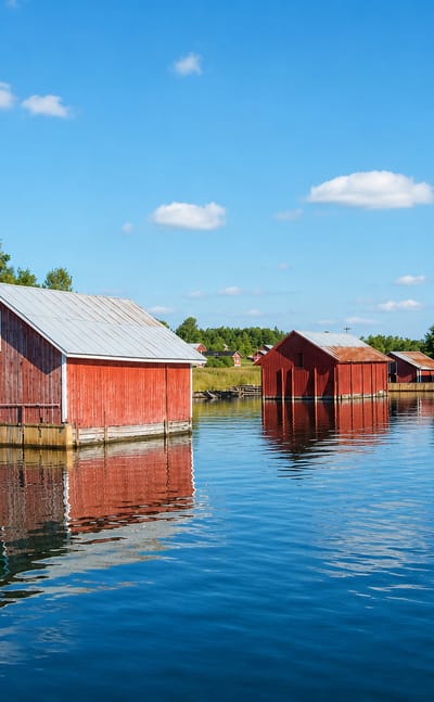 Red wooden boathouses reflected in calm archipelago waters under a blue summer sky in Houtskär, Finland.