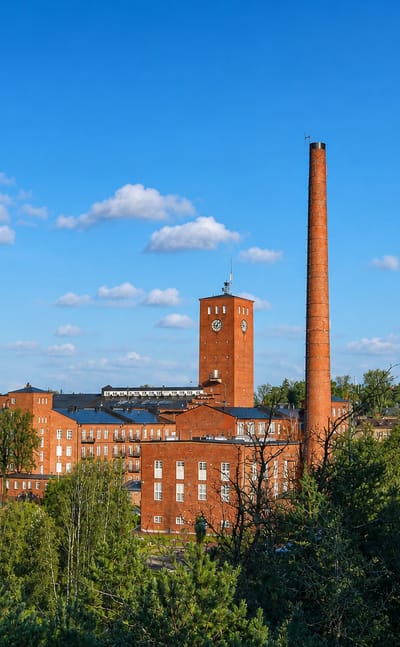 Red-brick industrial mill with a clock tower and tall chimney rising above green forest under a blue sky in Kaarina, Finland.