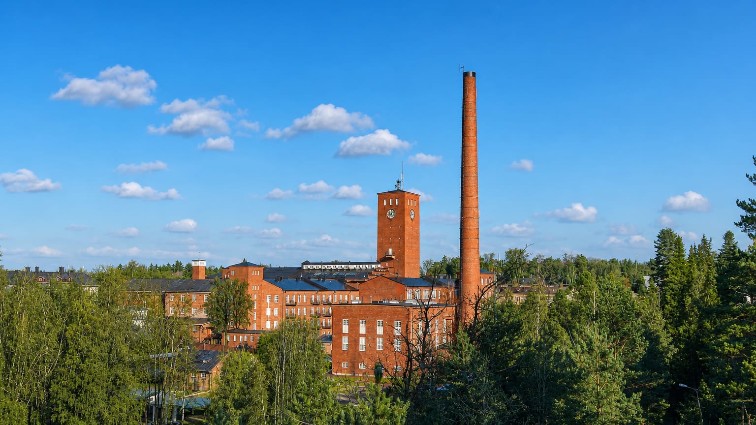 Red-brick industrial mill with a clock tower and tall chimney rising above green forest under a blue sky in Kaarina, Finland.