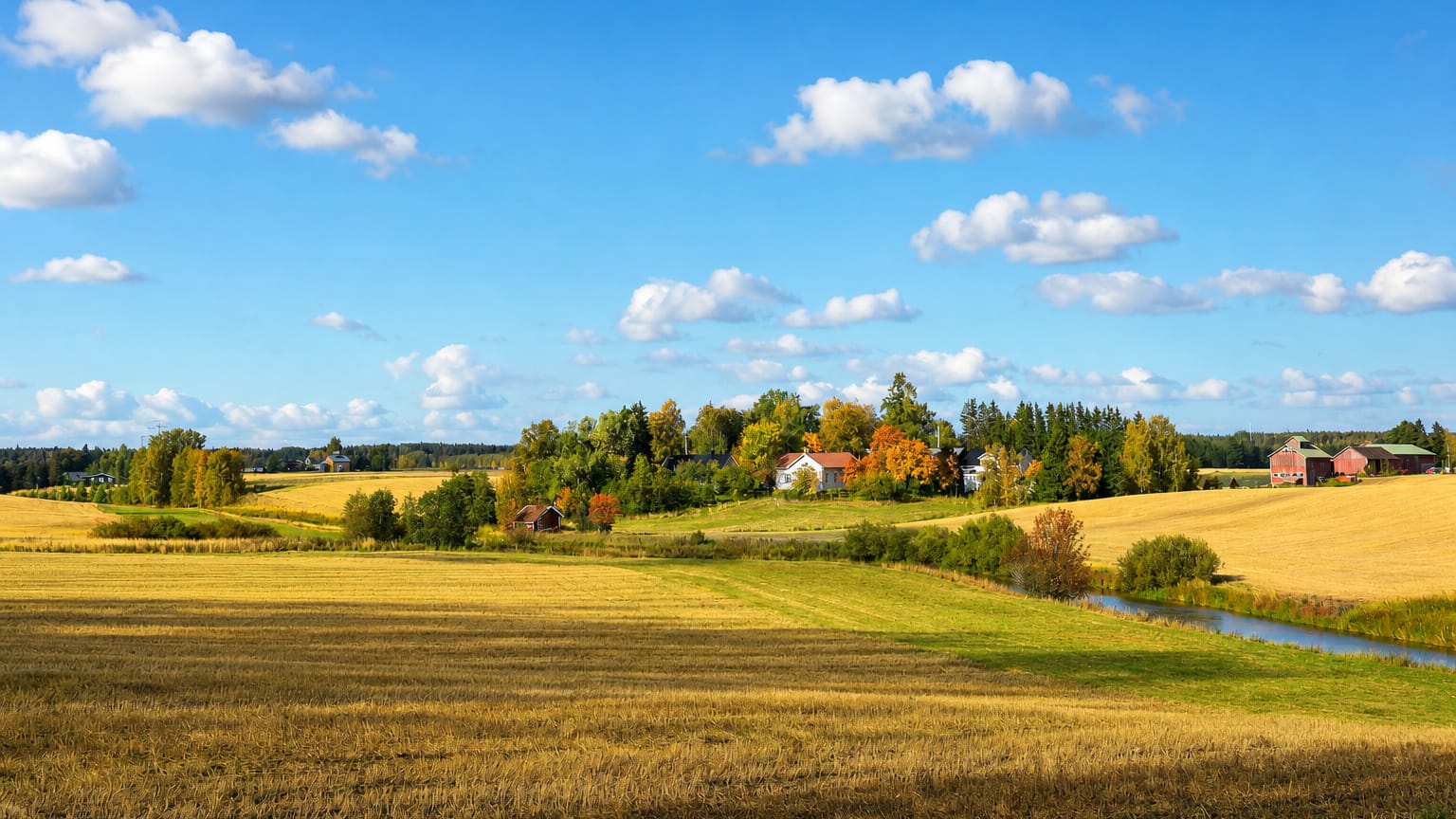 Rolling harvested farm fields, red barns and a farmhouse by a stream in Koski Tl, Finland.
