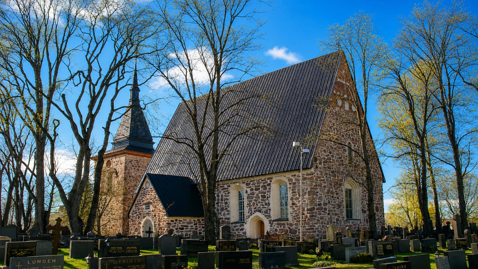 Medieval grey-stone church with a bell tower rising over a graveyard on a bright spring day in Laitila, Finland
