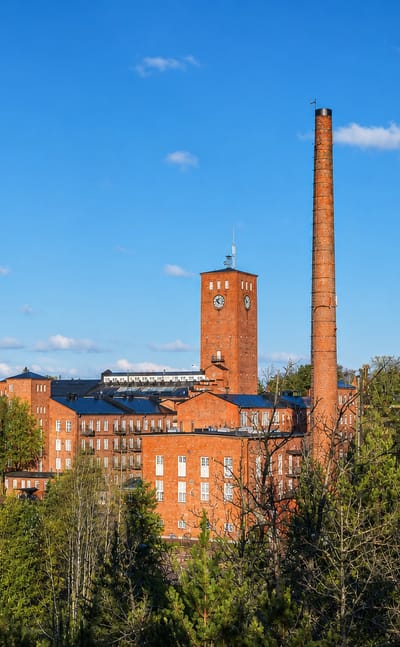 Historic red-brick mill complex with a tall chimney and clock tower rising above forest under a blue sky in Lieto, Finland