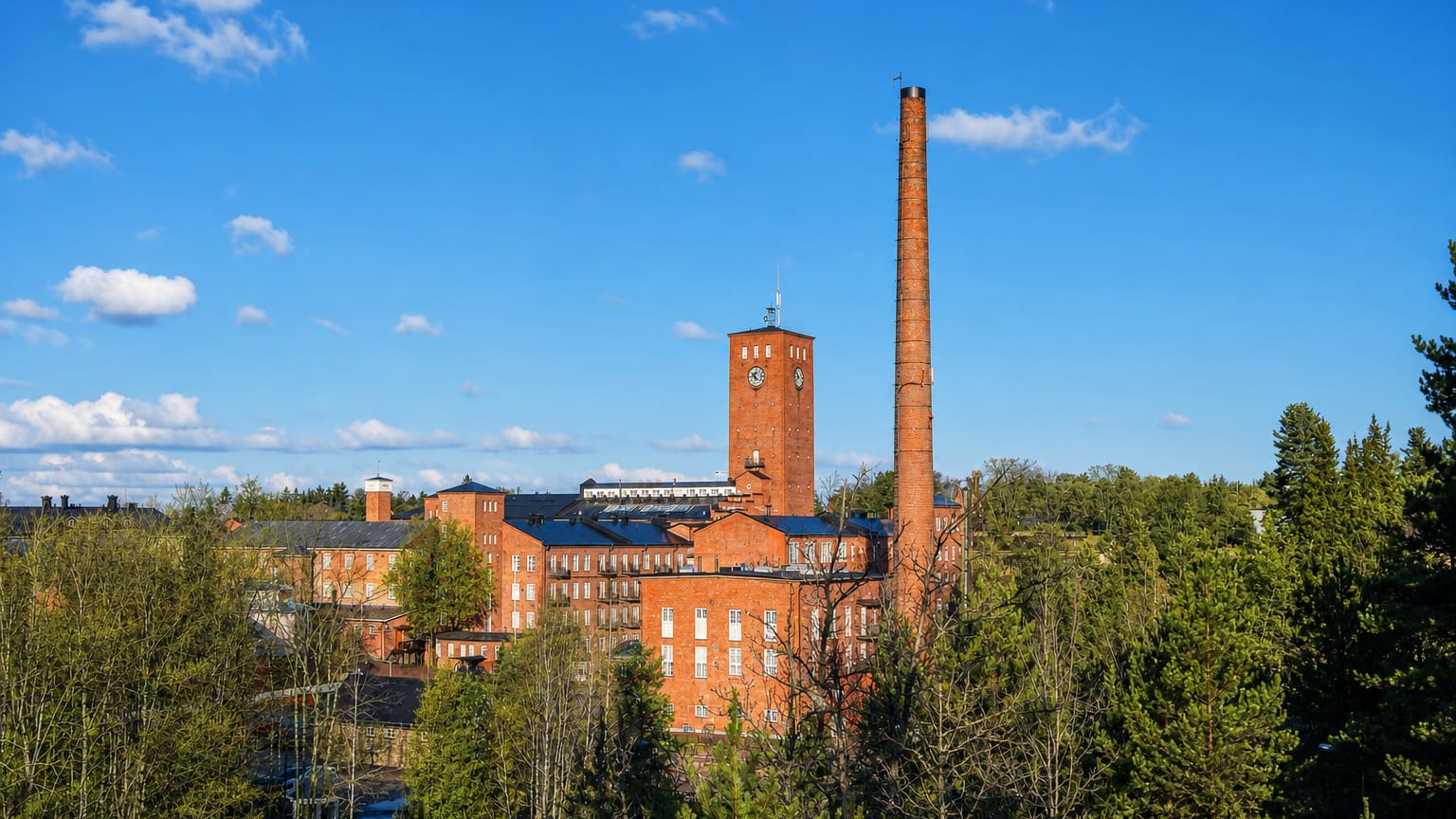 Historic red-brick mill complex with a tall chimney and clock tower rising above forest under a blue sky in Lieto, Finland