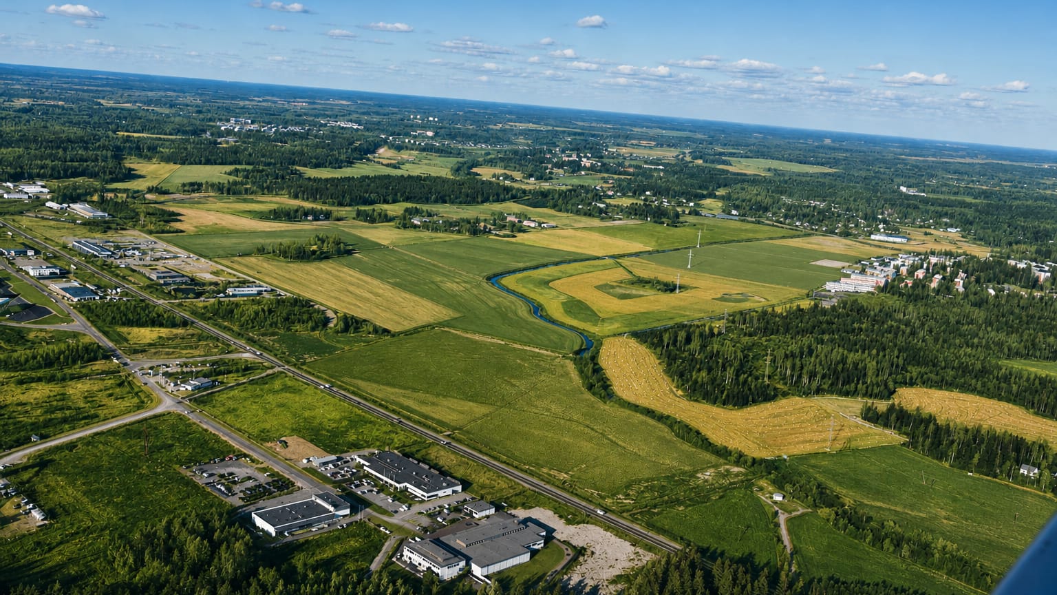 Aerial view over Marttila, Finland, with patchwork farmland, forest, a winding river and roadside buildings under a blue sky.