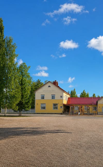 Yellow wooden buildings with red roofs and tall poplars beside a gravel square under a blue sky in Mellilä, Finland.