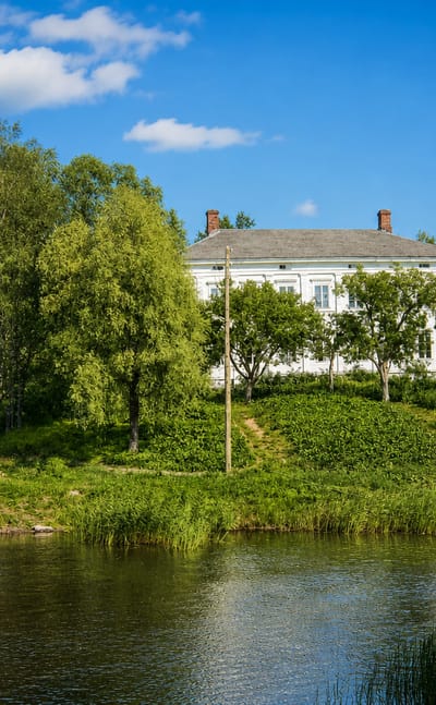 White wooden manor on a riverside hill with birches and an old stone footbridge in Mietoinen, Finland