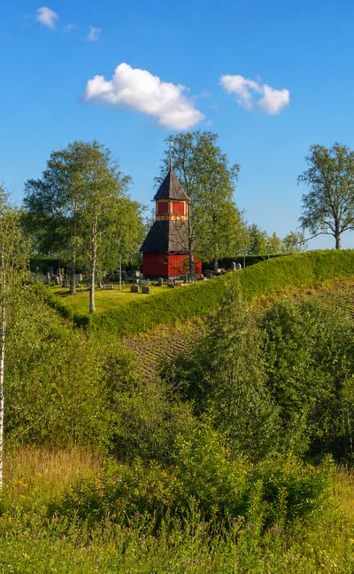 Red wooden church bell tower on a tree-lined hill above farm fields in Muurla, Finland, under a blue summer sky