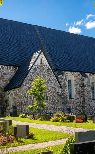 Grey medieval fieldstone church with a steep dark roof and graveyard in Mynamaki, Finland, on a sunny day.