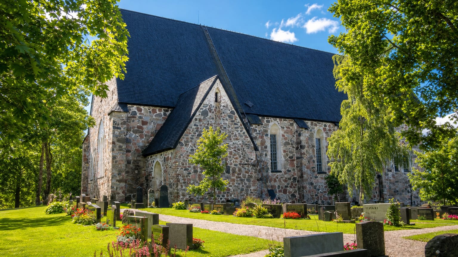 Grey medieval fieldstone church with a steep dark roof and graveyard in Mynamaki, Finland, on a sunny day.