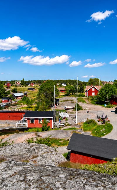 Nagu archipelago harbour with red wooden houses, boats at a jetty and granite shoreline under a bright summer sky.