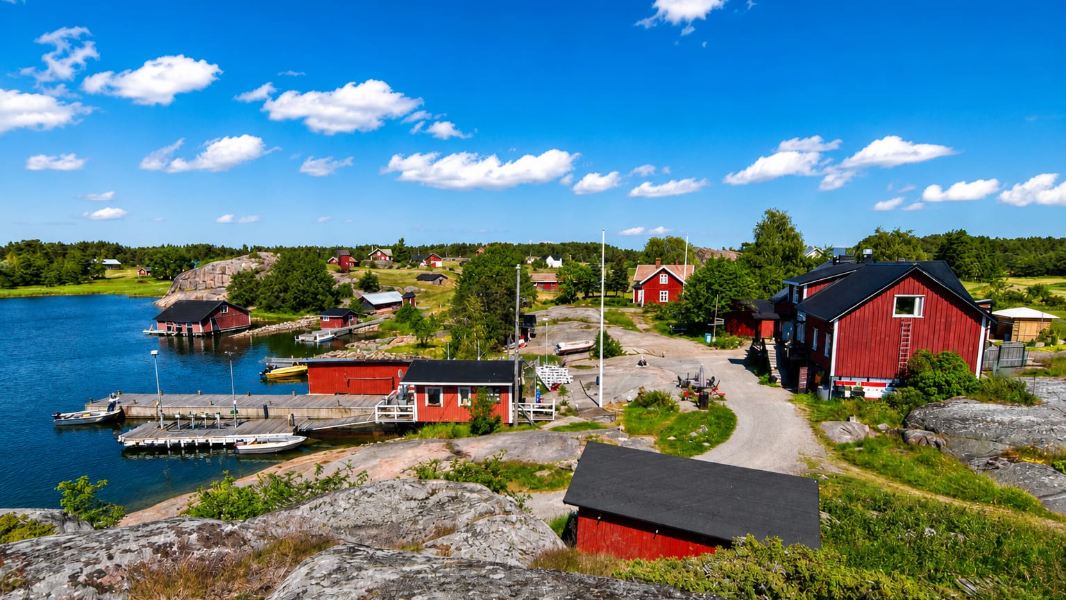 Nagu archipelago harbour with red wooden houses, boats at a jetty and granite shoreline under a bright summer sky.