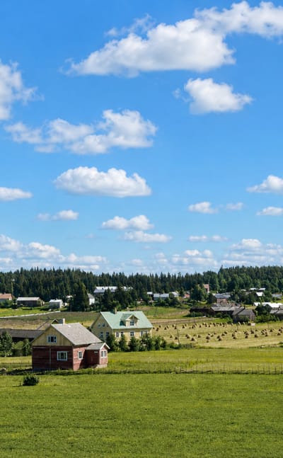 Rural Nousiainen, Finland: a red wooden farmhouse, hay bales and green fields below a conifer treeline and blue summer sky.