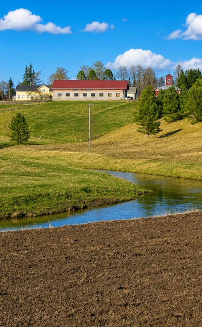 Rural Pertteli countryside in Finland with a plowed field, winding stream, green meadows and red-roofed farm buildings.