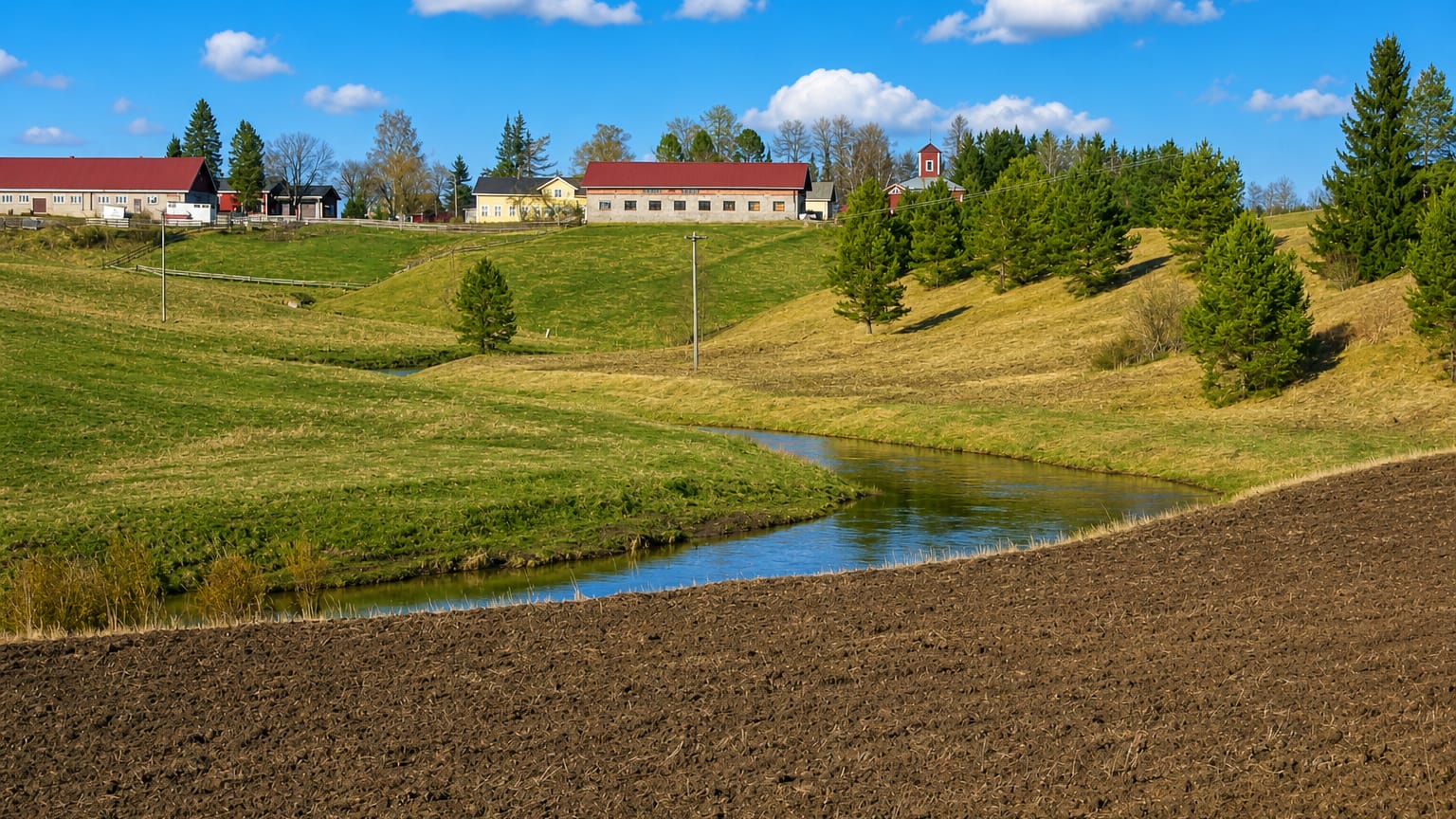 Rural Pertteli countryside in Finland with a plowed field, winding stream, green meadows and red-roofed farm buildings.
