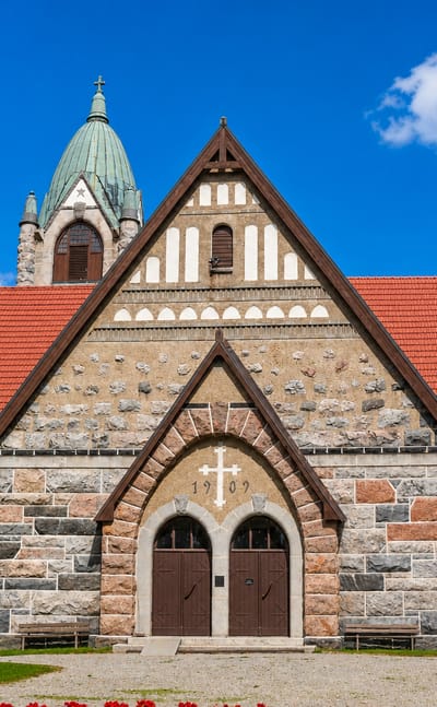 Grey stone church with a red gabled roof and green-domed tower in Pyhäranta, Finland, under a blue sky.