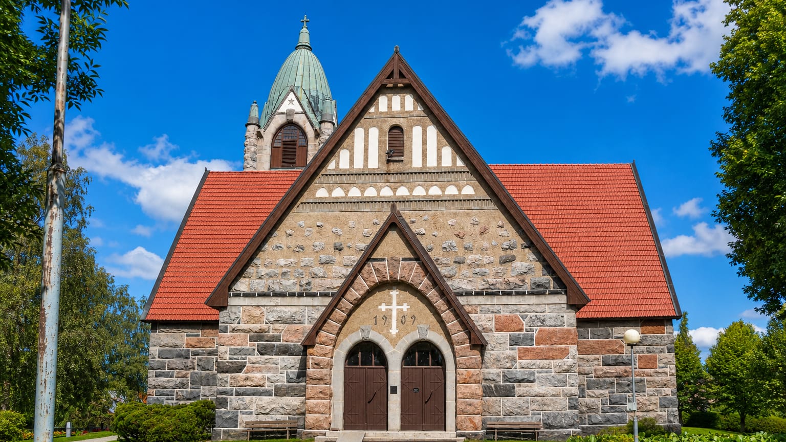 Grey stone church with a red gabled roof and green-domed tower in Pyhäranta, Finland, under a blue sky.