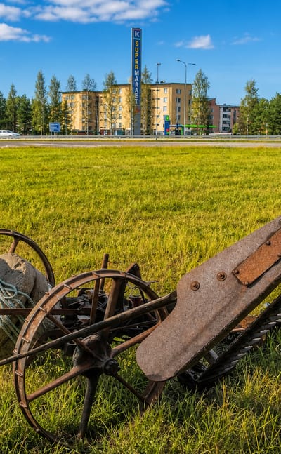 Antique rusty horse-drawn mower in a sunlit grassy field with apartment blocks and a road behind in Raisio, Finland.