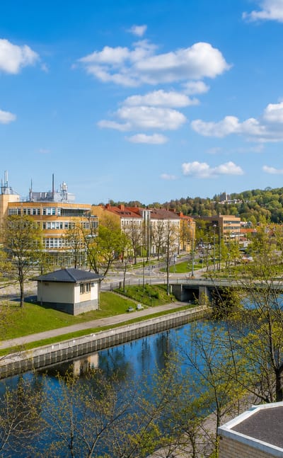 Riverside daytime townscape of Salo, Finland, with modern glass buildings, a bridge over the river and wooded hills under a blue sky.