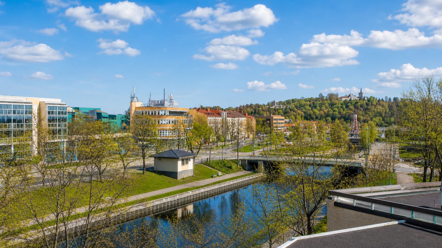 Riverside daytime townscape of Salo, Finland, with modern glass buildings, a bridge over the river and wooded hills under a blue sky.