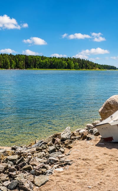 White rowing boat on Särkisalo's rocky, sandy shore beside a calm blue archipelago bay with forested islands beyond