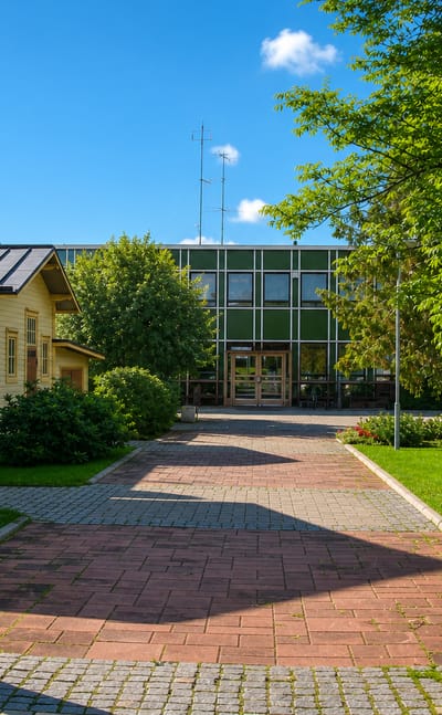 Brick-paved courtyard between a yellow timber building and a green-glass hall in Somero, Finland.