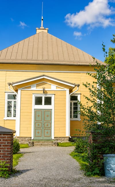 Suomusjärvi's yellow wooden church with a green door and pediment porch behind brick gateposts and trees.