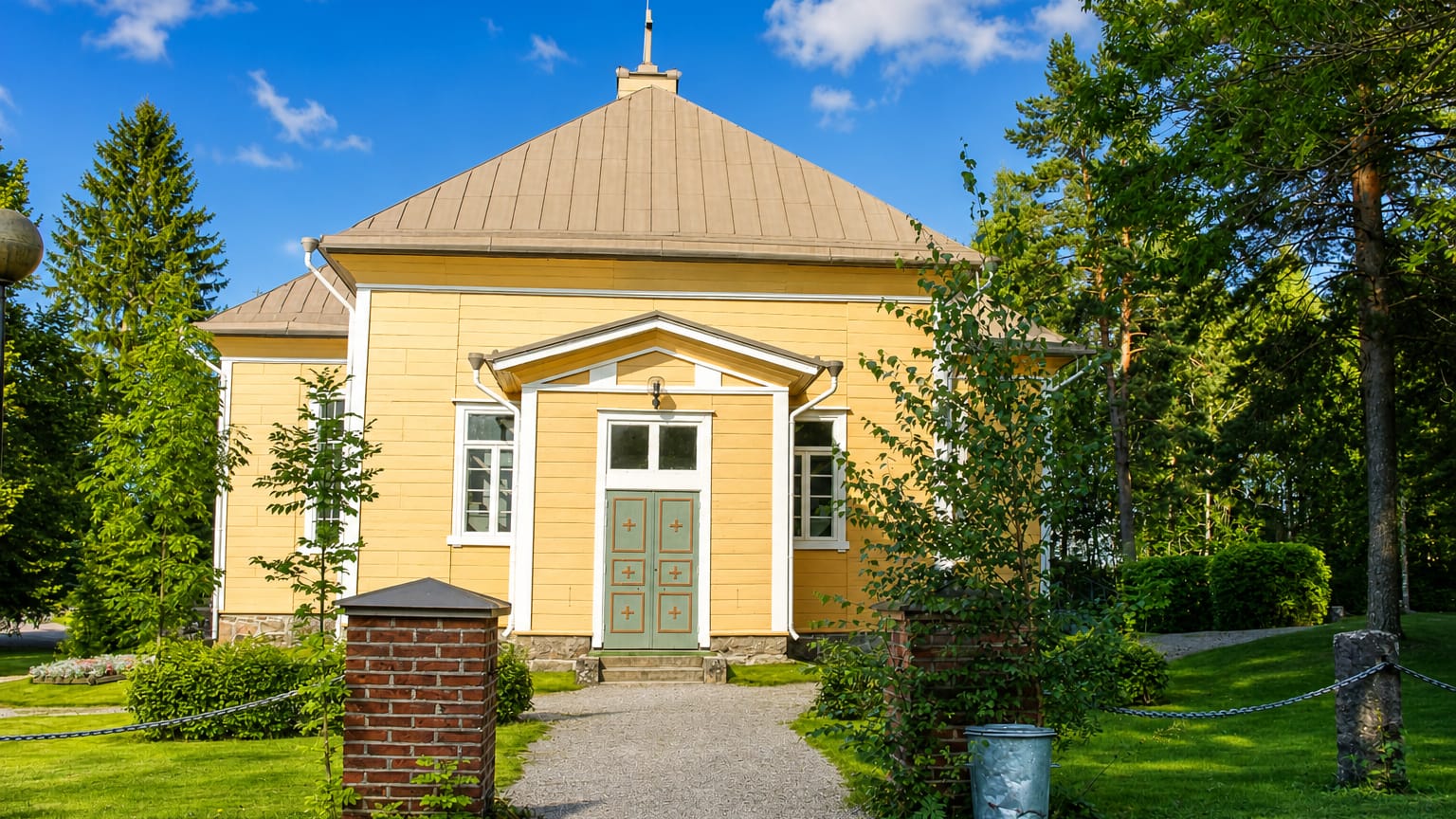 Suomusjärvi's yellow wooden church with a green door and pediment porch behind brick gateposts and trees.
