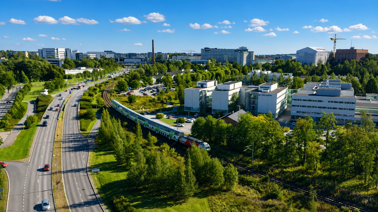 Aerial daytime view over I kaupunginosa, Turku: a highway, passing train, modern white buildings and green woodland under blue sky.