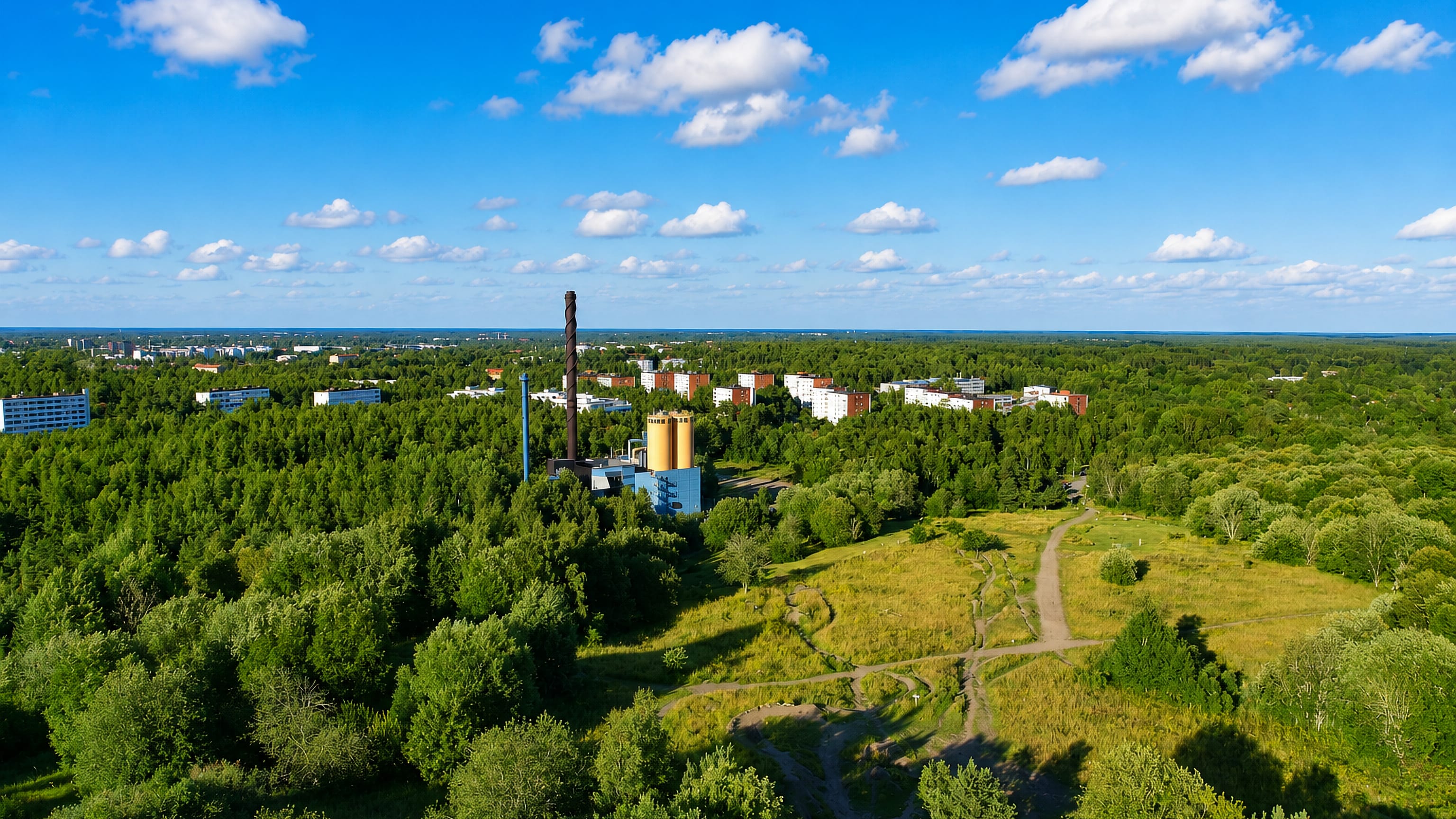 Aerial view of forest, meadow paths and a heating-plant chimney beside apartment blocks in Luolavuori, Turku.