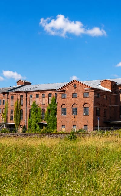Long red-brick industrial buildings along the railway in Raunistula, Turku, with a summer meadow in front.