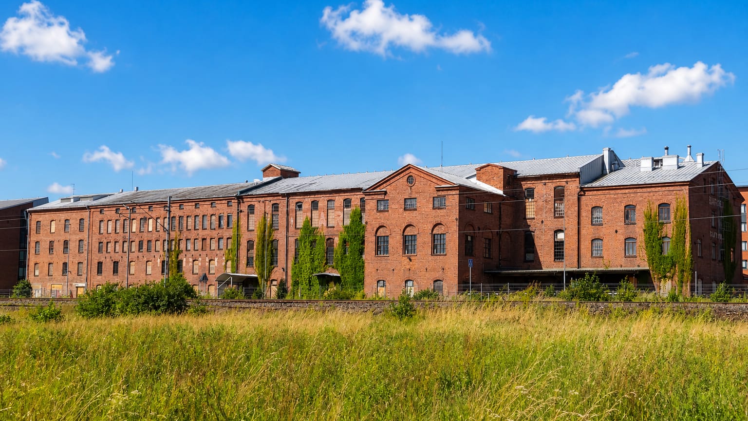 Long red-brick industrial buildings along the railway in Raunistula, Turku, with a summer meadow in front.