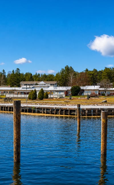 Velkua waterfront in Finland's archipelago, with low buildings on a wooded shore across calm water lined by weathered pier posts