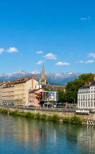 Grenoble quayside along the Isere with a church spire and snow-capped Alps rising behind the town.