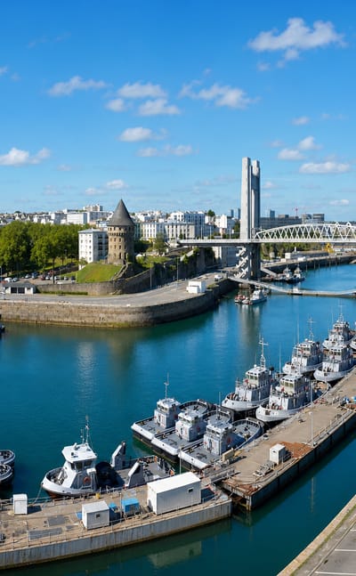 Brest's naval harbour with grey warships, the Recouvrance lift bridge and the round Tour Tanguy under blue sky.