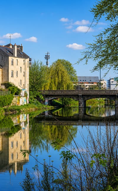 Stone bridge over a tree-lined canal in Rennes, France, with townhouses reflected in the calm water