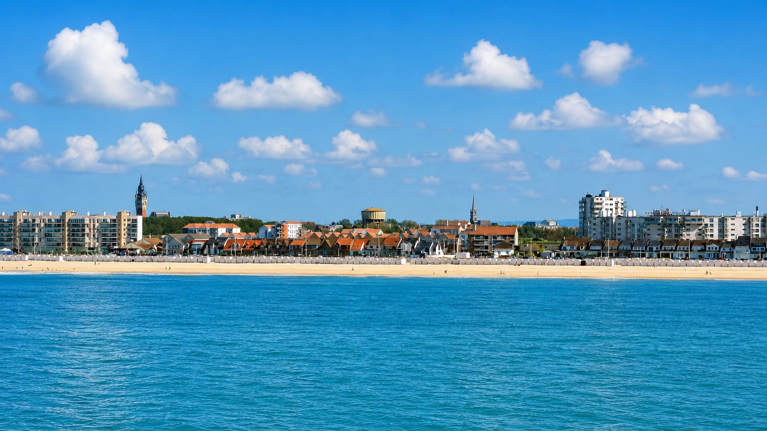 Calais seafront across the Channel, sandy beach backed by apartment blocks, a belfry spire and water tower.