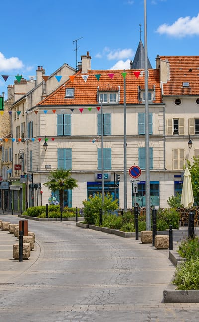 A sunny pedestrian street in old Nanterre with a pharmacy, cafe terrace, festive bunting and a small church spire