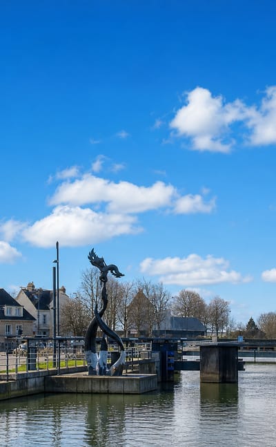 A canal lock and modern sculpture at Caen with stone Normandy houses and a distant church spire
