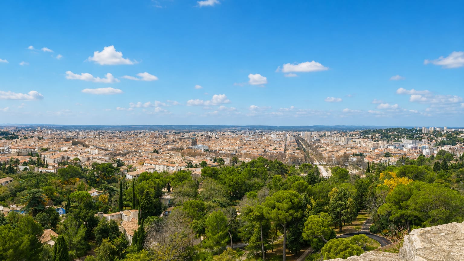 Elevated panorama over the terracotta rooftops of Nimes framed by Mediterranean pines and cypress under a blue sky