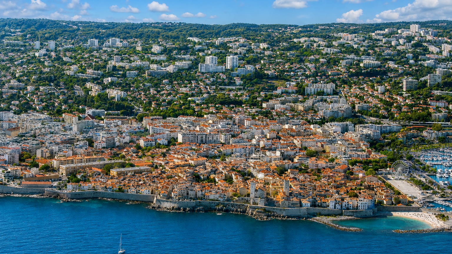 Aerial view of Antibes' walled old town and terracotta rooftops on a Mediterranean promontory beside its marina.