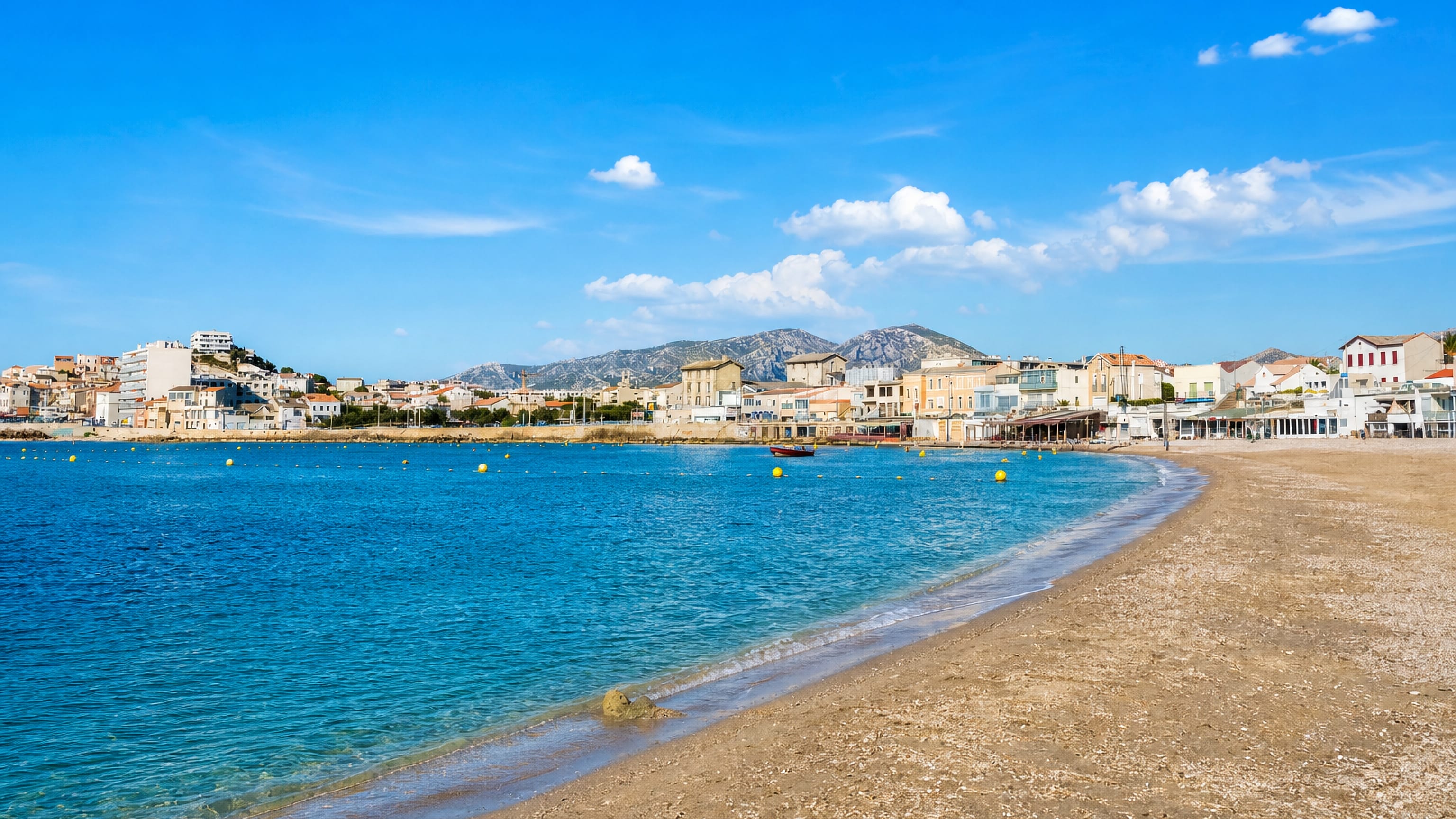 Sandy beach and turquoise water at La Pointe Rouge, Marseille, backed by hills and low houses.