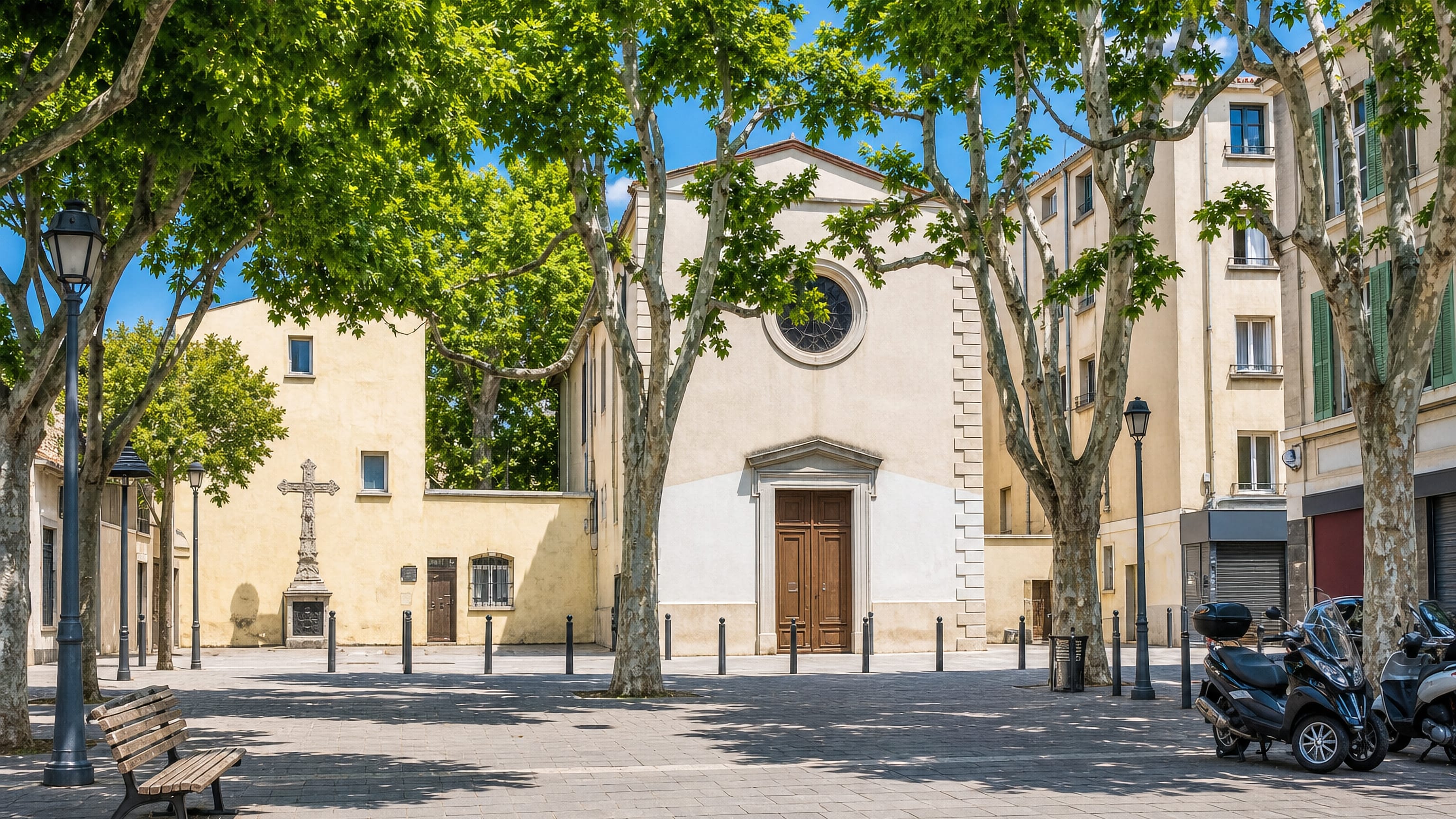 Shaded plane-tree square in Le Canet, Marseille, facing a cream-rendered church and a stone cross.